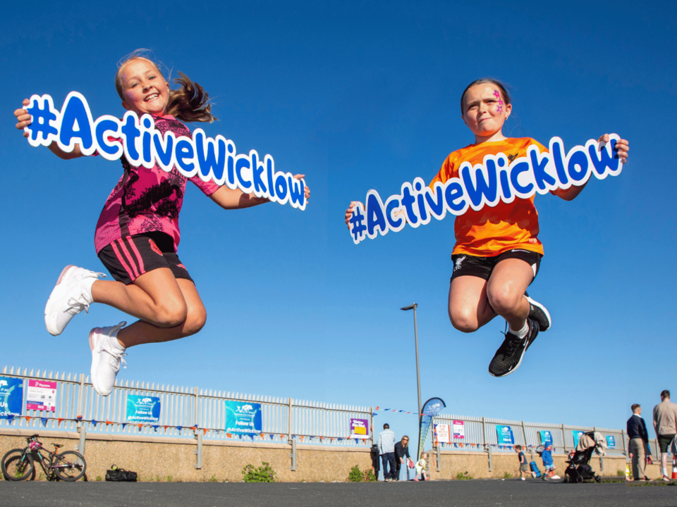 Two young girls jumping in the air, smiling and holding #ActiveWicklow signs against a clear blue sky at an outdoor event, with people, bicycles, and event posters visible in the background.