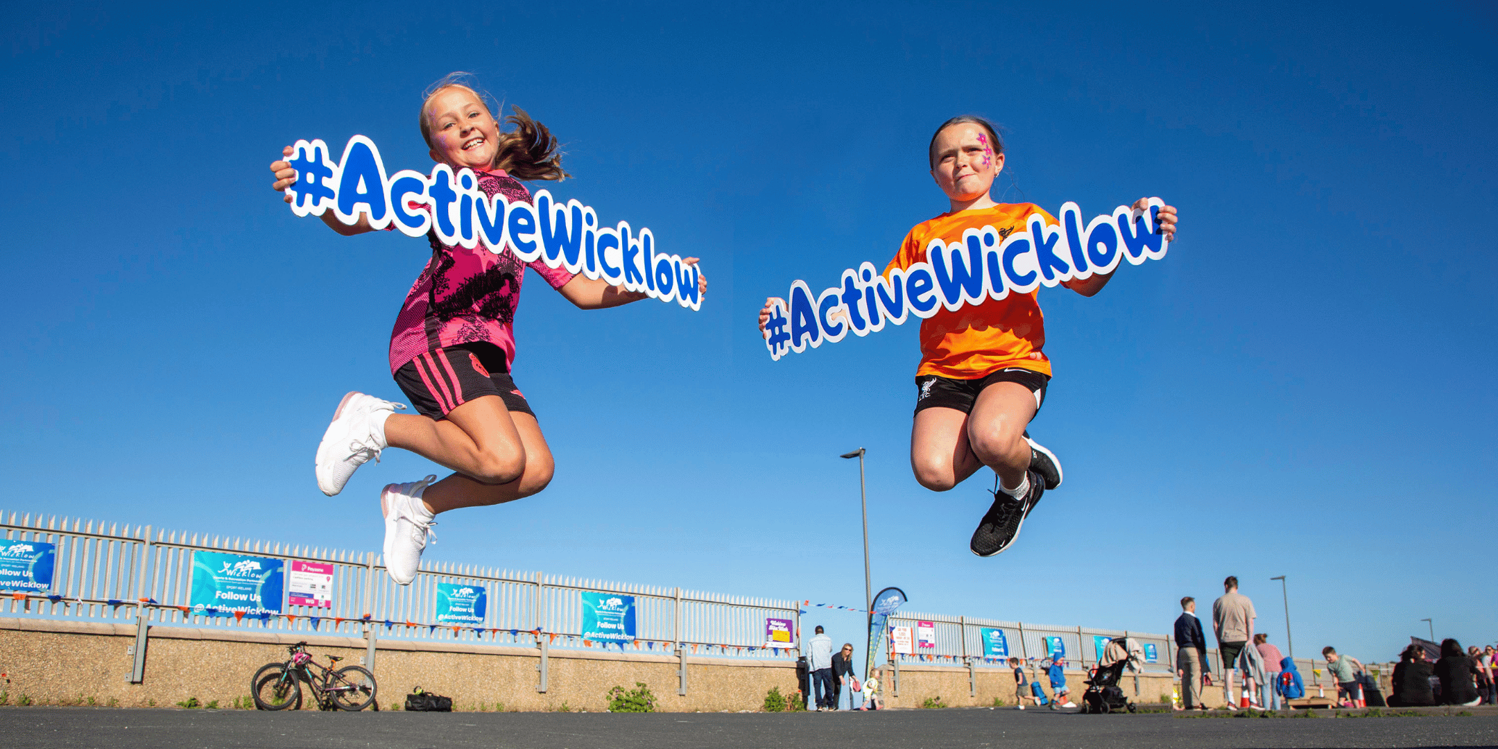 Two young girls jumping in the air, smiling and holding #ActiveWicklow signs against a clear blue sky at an outdoor event, with people, bicycles, and event posters visible in the background.